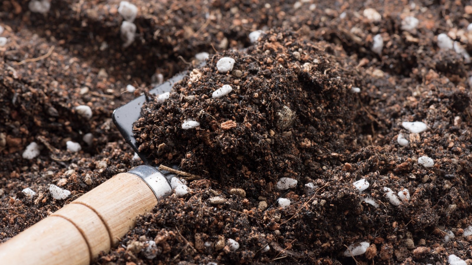 A shiny silver shovel is shown scooping up dark, moist material, with visible chunks and white particles mixed in, against a backdrop of scattered containers, emphasizing the precision and care in preparing the growing medium.