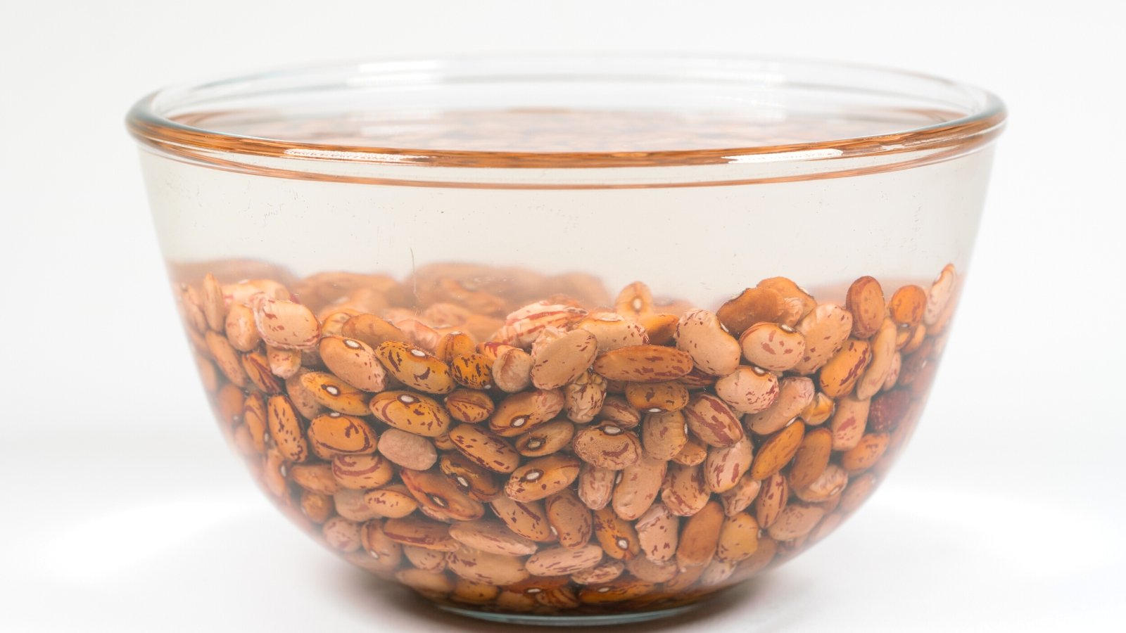 Close-up of bean seeds soaked in acid in a glass bowl on a white background. Rosecoco beans are oval-shaped and slightly flattened, with a smooth texture. They have a pale beige base color with vibrant pinkish-red streaks and spots covering their surface.