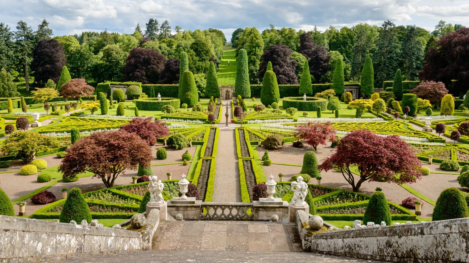 Drummond Castle stands majestically with its grand architecture overlooking impeccably designed formal gardens, characterized by intricate geometric patterns, manicured lawns, vibrant flower beds, and neatly trimmed topiary, all framed by a backdrop of lush greenery and ancient trees.