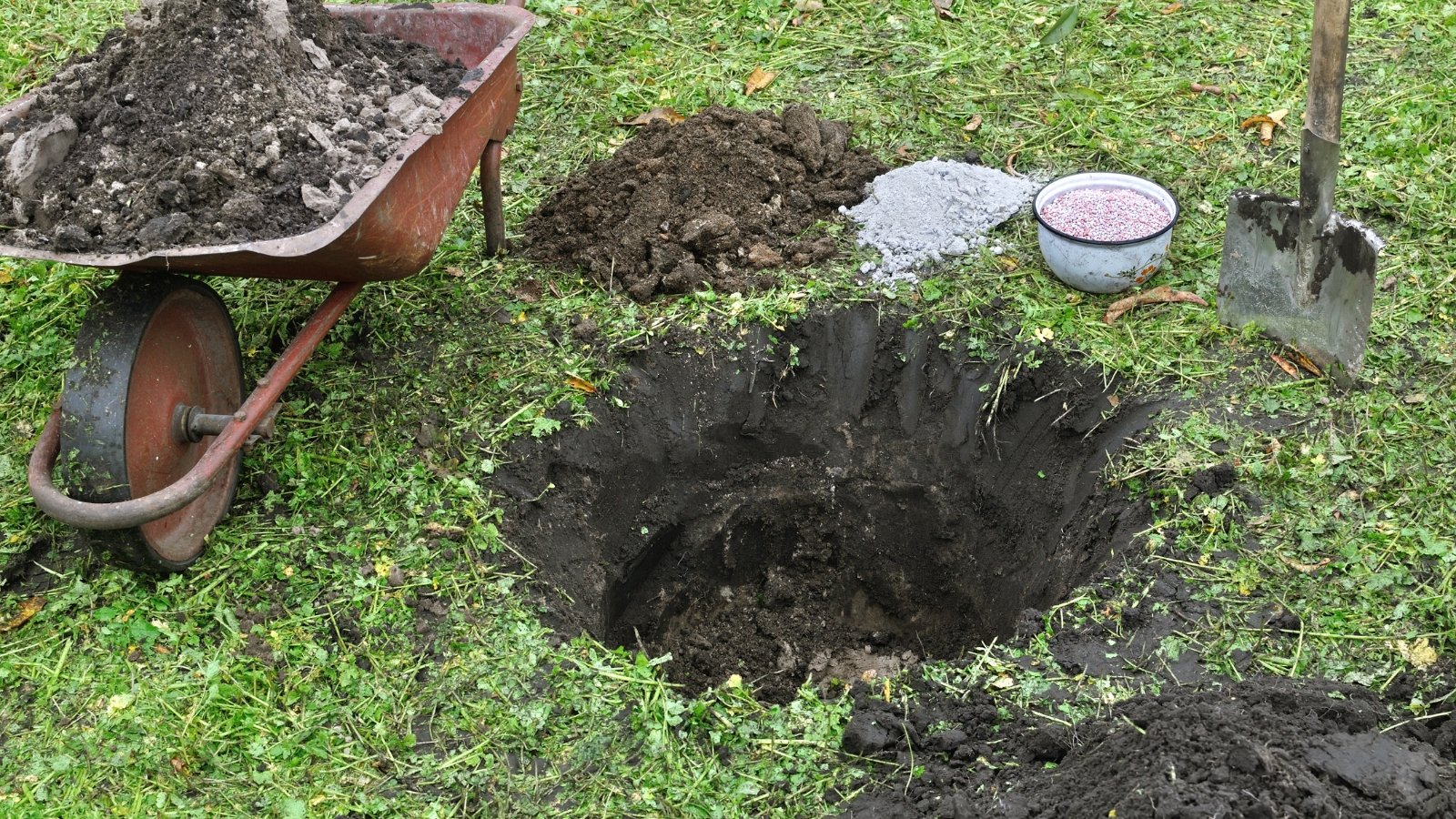 A deep hole with a pile of dark soil sits beside a wheelbarrow, shovel, and a round stepping stone.