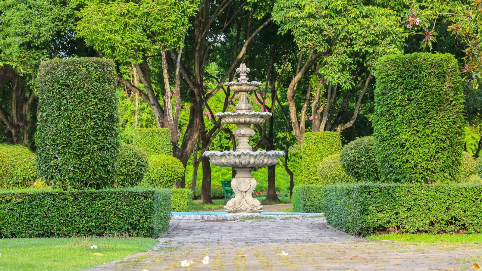 View of a formal garden landscape that features a central concrete architectural fountain, elegantly surrounded by neatly trimmed green box hedges, stately trees, and orderly walkways.