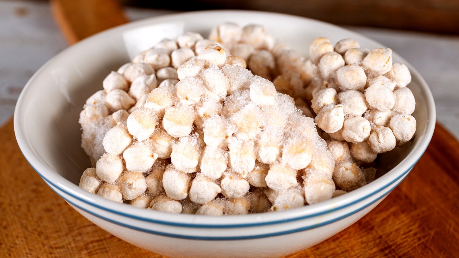 Close-up of frozen chickpeas seeds in a white ceramic bowl. Chickpea seeds are small, round, and slightly asymmetrical with a smooth surface. They have a beige to light tan color. Tiny ice pellets envelop the seeds and hold them together.