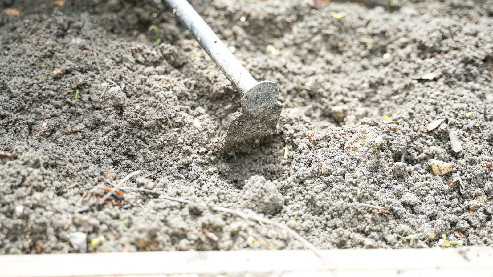 Sunlight casts shadows on a metal hoe resting against gray soil, hinting at the toil of gardening under the bright sky.