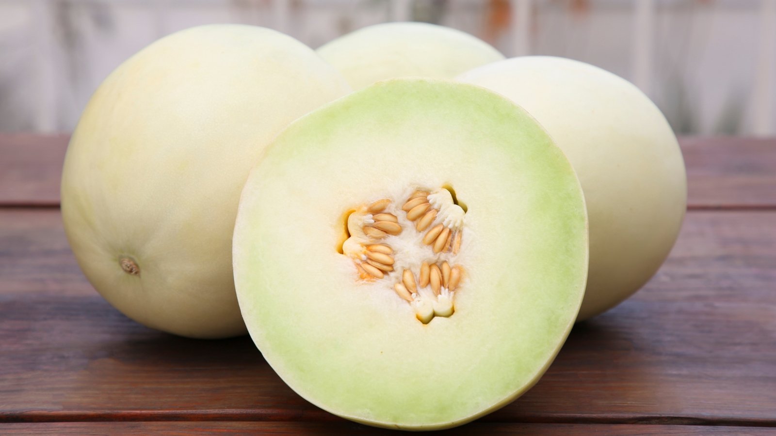 Three smooth, pale green melons and one cut open revealing light green flesh and clustered seeds rest on a wooden table.
