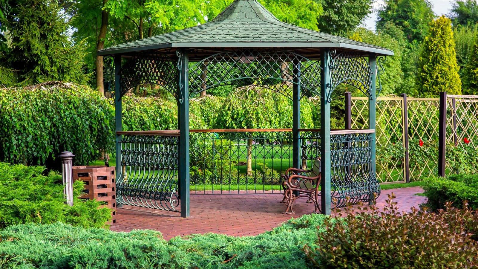 The iron gazebo, adorned with shingles and a bench, stands as a focal point in the formal garden, encircled by a variety of evergreens, lending an air of elegance and tranquility to the surroundings.
