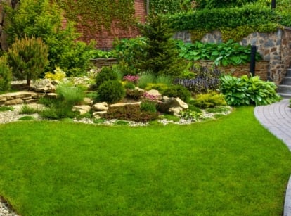 A vibrant green lawn framed by neatly arranged shrubs and flowers, bordered by a stone path leading to a cozy corner surrounded by dense greenery.