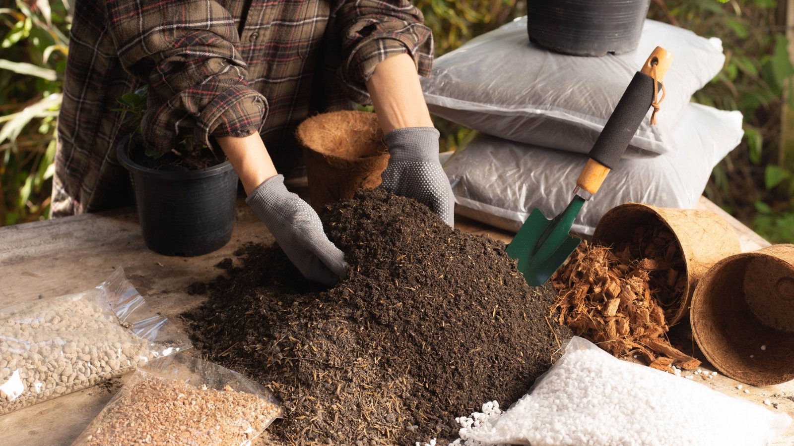 A pair of gloved hands work with a small green spade to mix a dark material on a wooden table, surrounded by planting containers and bags of materials, ready to create the ideal growing environment for new seedlings.