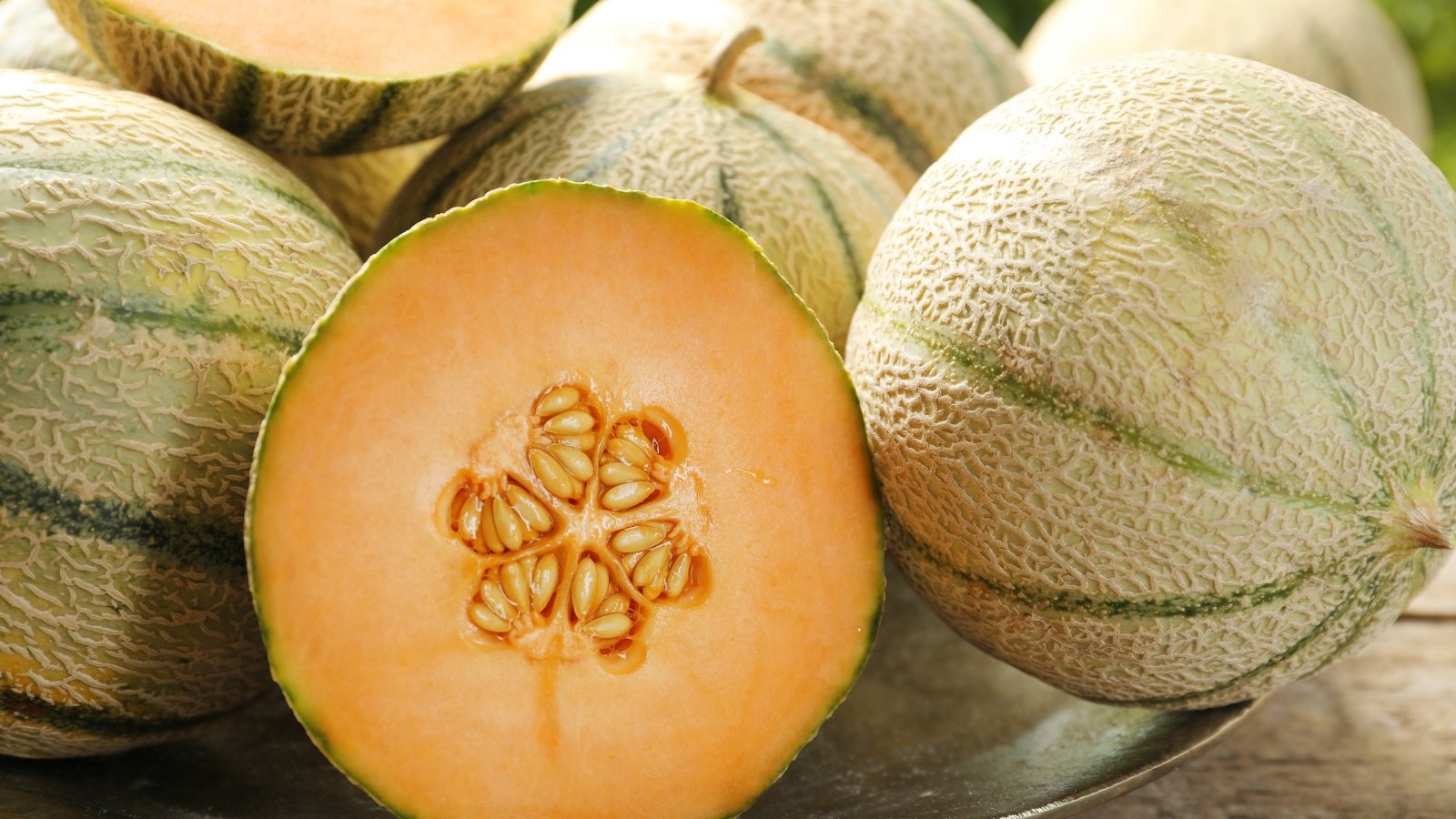 Several small, netted melons with tan skin rest on a table, one halved to display bright orange flesh and a seed-filled center.
