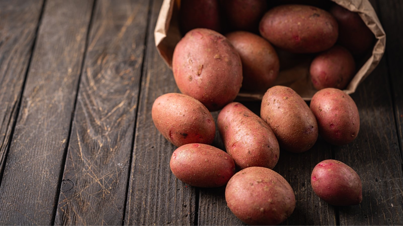A brown paper bag, slightly crumpled, sits atop a rustic wooden table, its edges curling gently. From its mouth spill red Cecile potatoes, each one a miniature marvel, promising earthy flavor and culinary adventure.