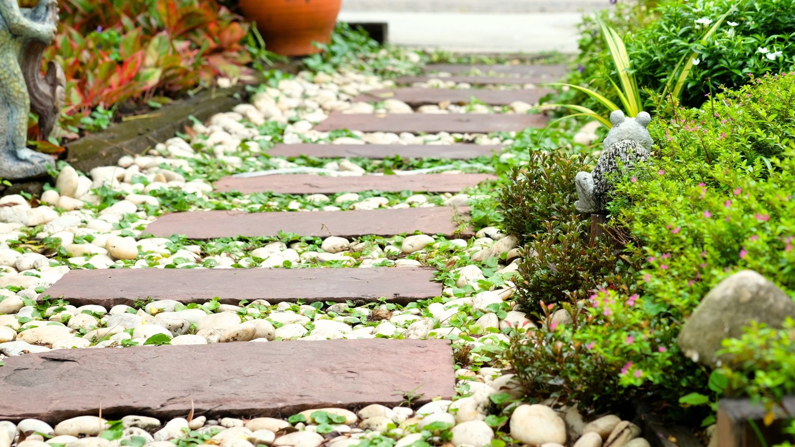 Close-up of a path made of white gravel and wooden planks laid parallel in the form of steps along a garden with various plants and decorative sculptures.
