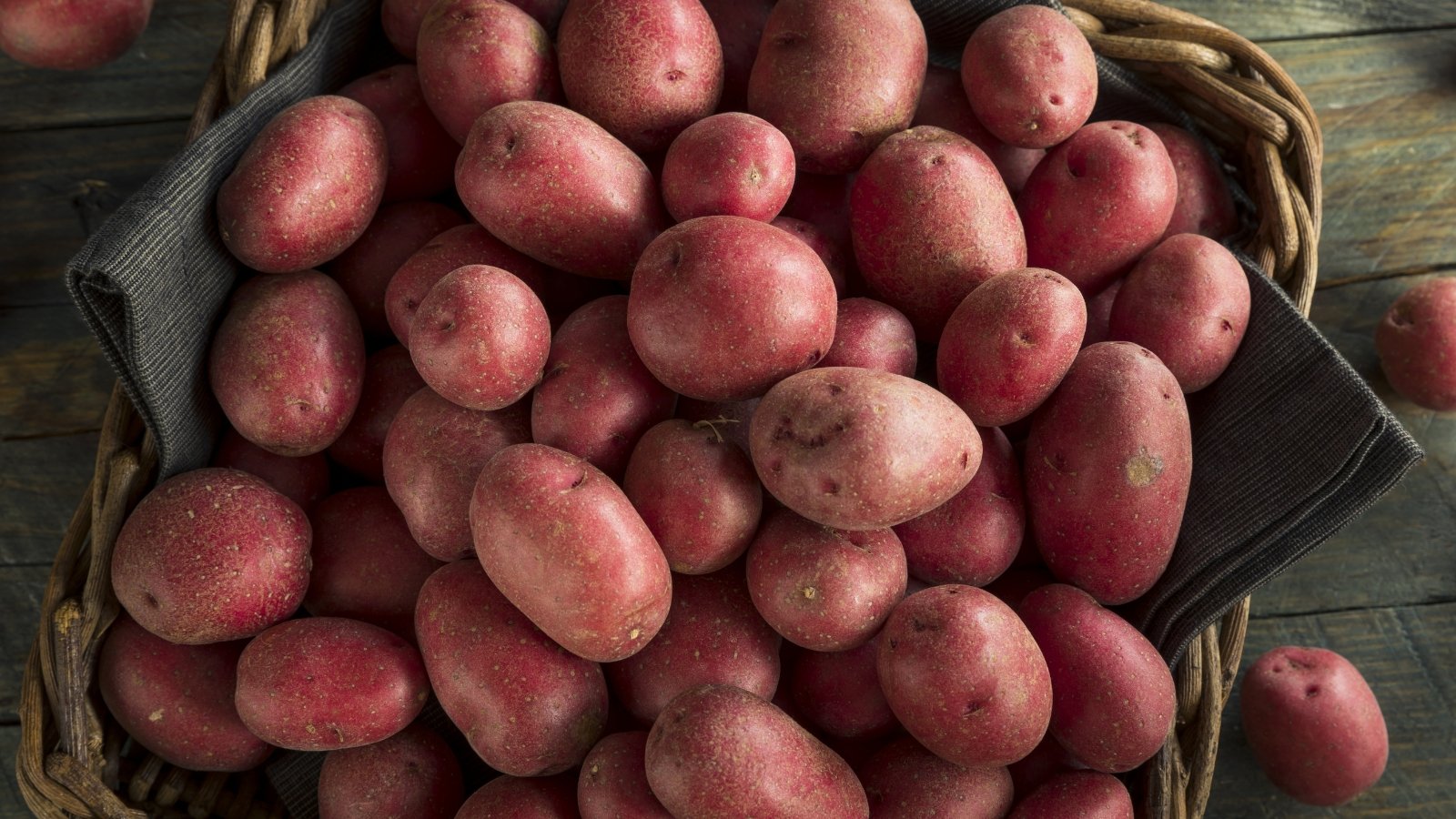 A woven basket holds vibrant red Sangre potatoes. Nestled on a wooden table, it presents a rustic charm. Some of the potatoes escape, adding a playful touch to the scene.