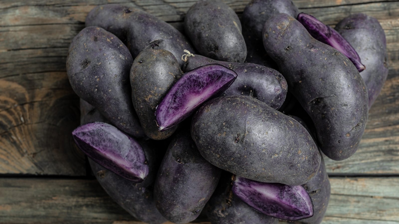 A bunch of Magic Molly potatoes sit atop a rustic wooden table, promising earthy delights. Among them, sliced potato rounds reveal a vibrant purple hue, tempting with their hidden colorful richness.