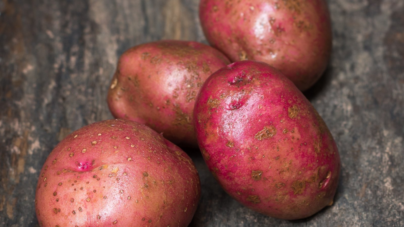 A close-up of Red Pontiac potatoes, arranged on a wooden surface, showcase their red skins under the warm glow of the sun. Each potato exudes a rustic charm, inviting the viewer to savor their earthy flavors and wholesome appeal.