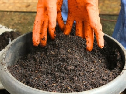 A gardener in orange gloves fills a gray container with soil, using a soil calculator to measure the right amount.