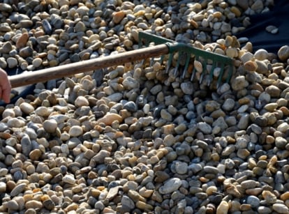 A hand using a rake to spread smooth, multicolored river rocks across a garden bed.